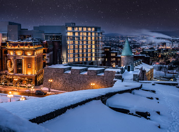 L’édifice moderne de l’hôtel parmi des bâtiments historiques sous la neige; le ciel est étoilé et la ville est illuminée au loin.