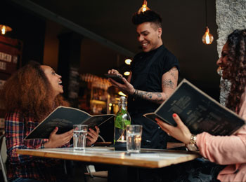 Deux femmes souriantes attablées à un restaurant, menu en main, s’adressant à la personne qui les sert.