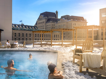 Quelques personnes se baignent dans une piscine extérieure; il y a de la neige au sol et des bâtiments historiques à l’horizon.
