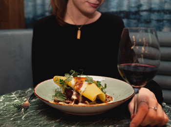 Assiette gastronomique de pâtes accompagnée d’un verre de vin rouge et présentée devant une femme en arrière-plan.