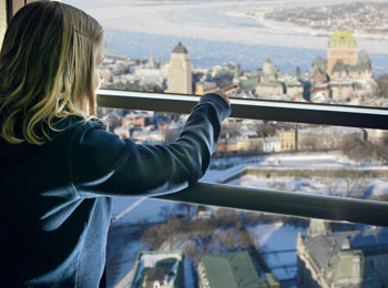 Une jeune fille se tient devant une fenêtre de l'Observatoire et elle pointe du doigt le Château Frontenac à l’horizon